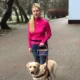Młoda kobieta w różowym swetrze trzyma smycz prowadzącą do jasnobrązowego psa przewodnika. Oboje znajdują się na zewnątrz, a w tle widać drzewa i ścieżkę. Kobieta jest osobą niewidomą, a pies nosi specjalną uprząż dla psów asystujących.