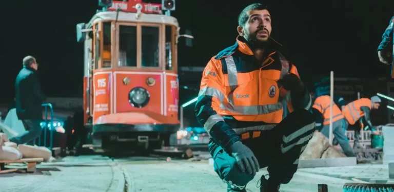 Na zdjęciu widać nocną scenę na placu budowy lub przy modernizacji torów tramwajowych. W centrum kadru klęczy młody mężczyzna w odblaskowej pomarańczowej kurtce roboczej z logo firmy na piersi. Jego twarz oświetla chłodne, sztuczne światło, a wyraz oczu zdradza skupienie i determinację. W tle widać klasyczny czerwony tramwaj o numerze 115, który zdaje się czekać na ponowne uruchomienie. Kilku innych pracowników w podobnych odblaskowych kurtkach jest zajętych pracą przy torowisku, otoczeni sprzętem i materiałami budowlanymi. Zdjęcie oddaje atmosferę ciężkiej pracy w nocy – chłodne powietrze, ostre światło reflektorów i intensywne skupienie pracowników. Można wyczuć poczucie odpowiedzialności oraz wytrwałość w realizacji zadania, niezależnie od trudnych warunków. Praca osoby niepełnosprawnej w nocy wymaga nie tylko fizycznego wysiłku, ale i determinacji, którą widać w spojrzeniu mężczyzny na pierwszym planie.