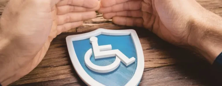 obrazek do artykułu - uprawnienia pracowników z orzeczeniem. Na zdjęciu widoczny jest symbol osoby na wózku inwalidzkim umieszczony na niebieskim tle w kształcie tarczy, sugerującym ochronę i bezpieczeństwo. Nad tarczą znajdują się dwie dłonie skierowane ku sobie, które wyglądają jakby chroniły symbol. Dłoń po lewej i po prawej stronie symbolu tworzą łuk, który nadaje całej scenie wrażenie opieki i wsparcia. Zdjęcie zostało wykonane na tle drewnianego blatu, co dodaje ciepła i naturalnego charakteru całości.