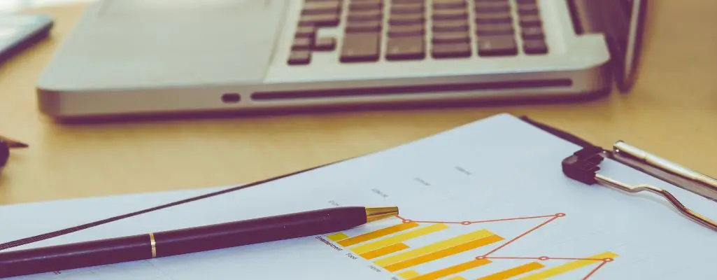 Wyjaśniamy, dlaczego uzyskanie ulgi PFRON jest skuteczną metodą na redukcję wpłaty PFRON 2 Na zdjęciu widać biurko z laptopem, długopisem i raportem zawierającym wykresy w odcieniach żółci i pomarańczy. Obraz oddaje atmosferę analizy finansowej i pracy biurowej. Sugeruje uzyskanie ulgi PFRON, podkreślając znaczenie dokumentacji i precyzji w formalnościach.