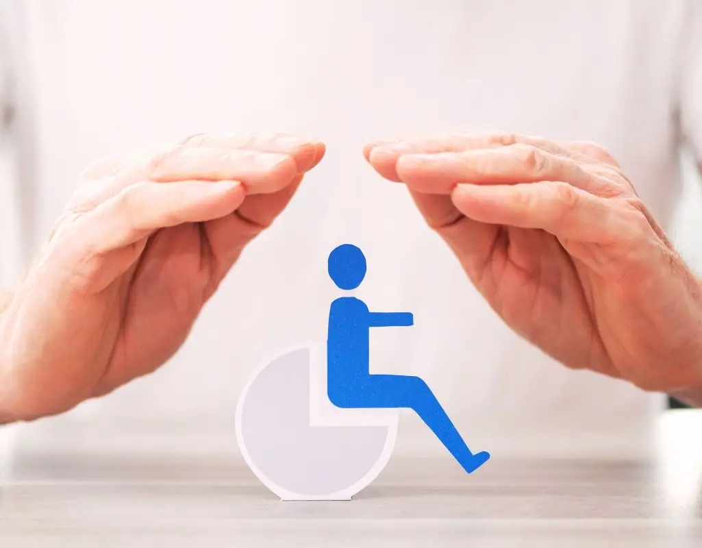 Pokazujemy wszystkie uprawnienia osób niepełnosprawnych, byś mógł z nich skorzystać 1 obrazek do artykułu - uprawnienia osób niepełnosprawnych. Na zdjęciu widoczne są dwie dłonie unoszące się nad niebieską ikoną symbolizującą osobę na wózku inwalidzkim, co symbolizuje ochronę praw i wsparcie dla osób niepełnosprawnych. Ikona osoby na wózku jest wykonana z prostych kształtów, a dłonie są złożone w geście ochronnym. Całość sugeruje ideę opieki i zabezpieczenia praw osób z niepełnosprawnością.