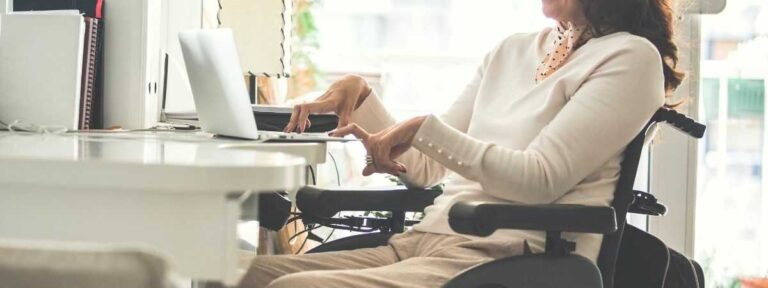 obrazek do artykułu - Przywileje osób niepełnosprawnych w pracy. Na zdjęciu widzimy osobę na wózku inwalidzkim, siedzącą przy biurku, pracującą na laptopie. Osoba jest ubrana w jasny sweter i beżowe spodnie, co tworzy stonowany, elegancki styl. Ręce są wyciągnięte w kierunku laptopa, a postawa sugeruje skupienie na wykonywanych czynnościach. W tle widoczne są jasne wnętrza, co nadaje scenie przyjazny i profesjonalny charakter. Zdjęcie sugeruje tematykę związaną z pracą lub nauką osób z niepełnosprawnościami.