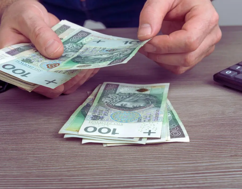 obrazek do artykułu - Jakie są warunki otrzymania dofinansowania wynagrodzeń? Na zdjęciu widzimy mężczyznę trzymającego w dłoni banknoty o nominale 100 złotych, charakterystyczne dla polskiej waluty. Na stole przed nim leży stos podobnych banknotów, a obok znajduje się kalkulator. Obrazek symbolizuje liczenie pieniędzy lub zarządzanie finansami. Można odczuć atmosferę związania z oszczędnością, planowaniem wydatków lub przygotowaniem do ważnej decyzji finansowej. Przedstawione ręce w działaniu podkreślają praktyczny aspekt codzienności i odpowiedzialności finansowej.
