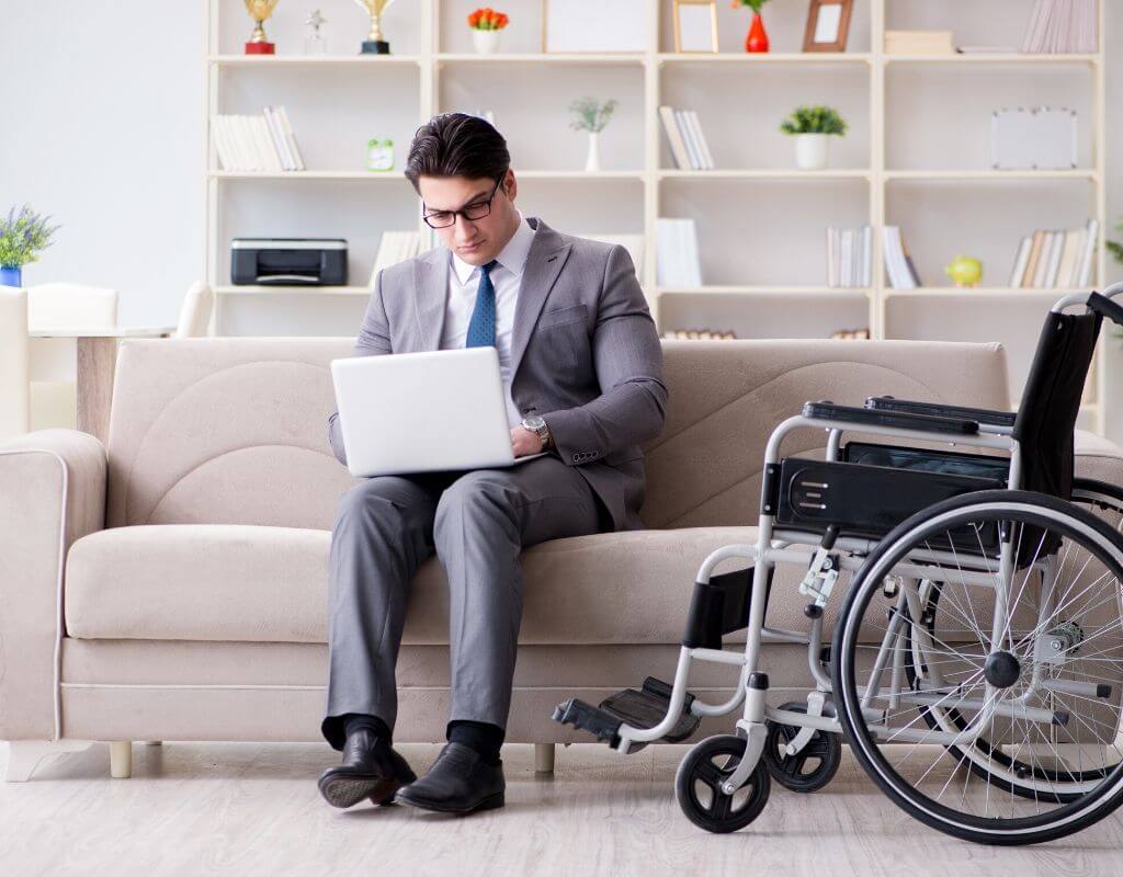 Gdzie szukać pracy zdalnej, by to było skuteczne? 1 Obrazek wyróżniający do artykułu - gdzie szukać pracy zdalnej? Zdjęcie przedstawia mężczyznę siedzącego na kanapie i pracującego na laptopie w swoim domu, obok niego stoi wózek inwalidzki.