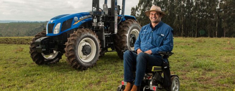 Na czym polega refundacja składek KRUS z PFRON? Zasady 2026, dla kogo, jak złożyć wniosek 7 Zdjęcie w artykule - PFRON refundacja składek KRUS. Pokazuje uśmiechniętego mężczyznę w niebieskiej roboczej koszuli, siedzącego na elektrycznym wózku inwalidzkim na tle zielonego pola i dużego niebieskiego traktora przystosowanego do obsługi przez osobę niepełnosprawną. Zdjęcie oddaje poczucie niezależności i dumy z pracy w rolnictwie, a także potencjał technologii wspierającej osoby z niepełnosprawnościami.