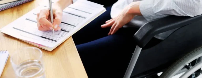 Obrazek wyróżniający do artykułu - wsparcie dla pracodawców zatrudniających niepełnosprawnych. Na zdjęciu znajduje się osoba siedząca na wózku inwalidzkim, pisząca coś na kartkach papieru leżących na stole. Osoba trzyma w prawej ręce długopis i wypełnia dokumenty, które są rozłożone przed nią. Na stole, obok dokumentów, znajduje się również szklanka z wodą. Atmosfera na zdjęciu sugeruje pracę biurową lub wypełnianie formalności.