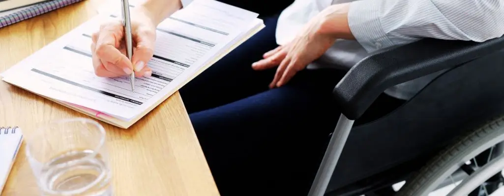 Przedstawiamy kompleksowe wsparcie dla pracodawców zatrudniających niepełnosprawnych, abyś wykorzystał wszystkie możliwości 1 Obrazek wyróżniający do artykułu - wsparcie dla pracodawców zatrudniających niepełnosprawnych. Na zdjęciu znajduje się osoba siedząca na wózku inwalidzkim, pisząca coś na kartkach papieru leżących na stole. Osoba trzyma w prawej ręce długopis i wypełnia dokumenty, które są rozłożone przed nią. Na stole, obok dokumentów, znajduje się również szklanka z wodą. Atmosfera na zdjęciu sugeruje pracę biurową lub wypełnianie formalności.