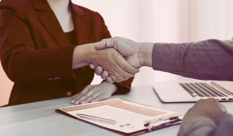 Na zdjęciu uchwycono moment uścisku dłoni dwóch osób przy biurku – jedna z nich ma na sobie elegancką marynarkę, druga koszulę. Na blacie widać wypełniony formularz, długopis i otwarty laptop. To symboliczna scena kończąca rozmowę kwalifikacyjną, pełna profesjonalizmu, szacunku i nadziei na dobrą współpracę. Zdjęcie świetnie oddaje atmosferę, jaka powinna towarzyszyć procesowi, jakim jest rekrutacja osób niepełnosprawnych – zaufanie, równe traktowanie i poczucie, że każda ze stron została wysłuchana i doceniona.