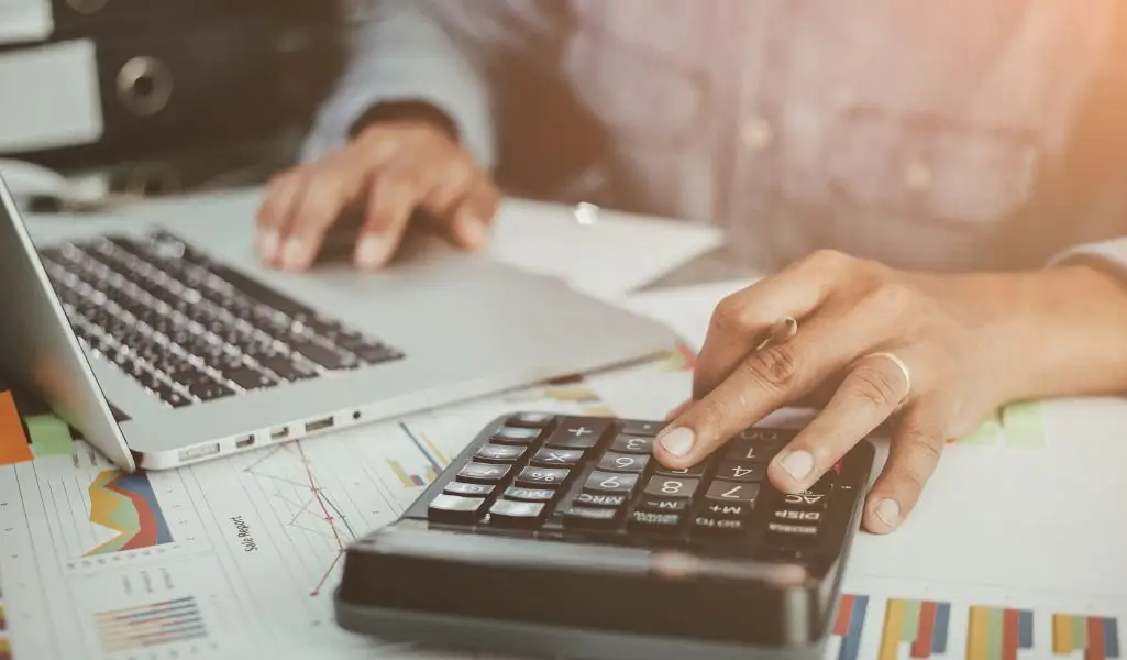 Jak otrzymać dofinansowanie na przystosowanie stanowiska pracy pracownika niepełnosprawnego w 5 krokach 2 obrazek wyróżniający w artykule - Środki na przystosowanie stanowiska pracy. Zdjęciu osoba pracuje na laptopie oraz kalkulatorze. Pod urządzeniami liczne dokumenty.