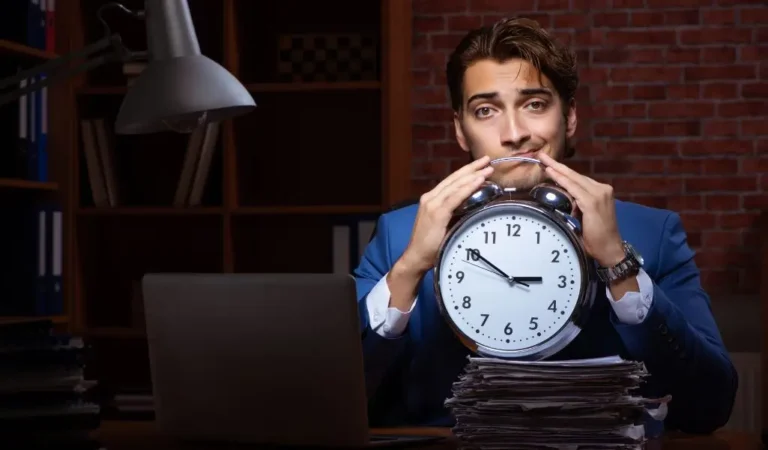 Obrazek wyróżniający w artykule - nadgodziny pracownika niepełnosprawnego. Na zdjęciu mężczyzna w ciemnym pomieszczeniu trzyma dłonie w ochronnym geście ponad tradycyjnym budzikiem. Zdjęcie sugeruje późną porę i zmęczenie pracownika, który pracuje w nadgodzinach.