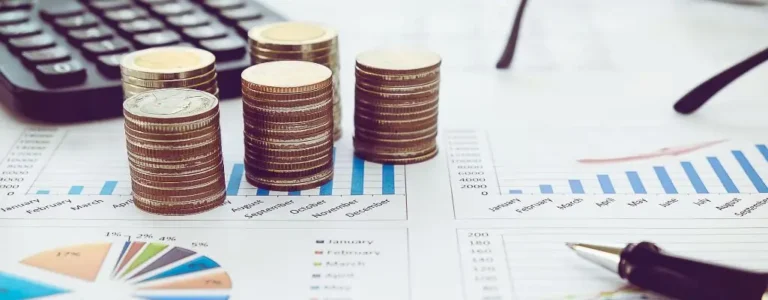 Obrazek do artykułu - dofinansowanie wynagrodzenia PFRON od 2020 roku. Na zdjęciu widzimy kilka niewysokich, równych stosów monet ułożonych na wydrukowanych wykresach i tabelach finansowych. W tle znajduje się kalkulator oraz długopis. Obraz przywodzi na myśl skupienie na analizie finansowej, poczucie kontroli nad budżetem oraz dążenie do stabilności i bezpieczeństwa w zarządzaniu pieniędzmi.