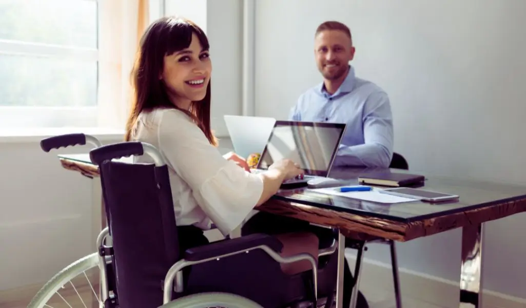 Jak zatrudnić pracownika niepełnosprawnego zgodnie z wymogami prawa i korzyścią dla Twojej firmy? 5 Na zdjęciu znajduje się uśmiechnięta kobieta na wózku inwalidzkim, siedząca przy drewnianym biurku w nowoczesnym, dobrze oświetlonym biurze. Ma na sobie jasną bluzkę i pracuje na laptopie, odwracając się w stronę obiektywu z promiennym uśmiechem. Po drugiej stronie biurka siedzi mężczyzna w koszuli, który również się uśmiecha. Na stole znajdują się notatniki, długopisy i dokumenty, co sugeruje, że jest to środowisko pracy. Zdjęcie oddaje atmosferę profesjonalizmu, równości i integracji, pokazując, że osoby z niepełnosprawnościami mogą z powodzeniem rozwijać się zawodowo. Idealnie ilustruje temat jak zatrudnić pracownika niepełnosprawnego, podkreślając znaczenie otwartości, dostępności i przyjaznego miejsca pracy.