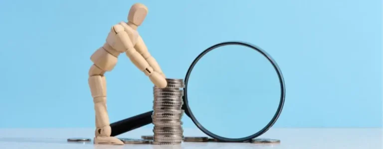 Obrazek wyróżniający w artykule - Wysokość dofinansowania wynagrodzeń PFRON dla firmy. Zdjęcie przedstawia manekina, symbolizującego człowieka, który ułożył monety w stos obok lupy. Manekin jest pochylony nad monetami, co może sugerować analizę lub dokładne sprawdzenie stanu finansowego. Zdjęcie to może oddawać uczucie dokładności, precyzji, a także niezbędnej ostrożności przy zarządzaniu finansami.