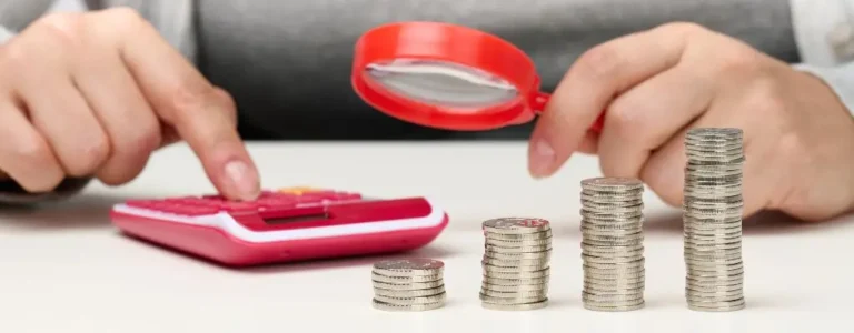 Obrazek wyróżniający w artykule - Dofinansowanie wynagrodzeń. Przedstawia ręce osoby używającej kalkulatora oraz ułożone obok stosy monet różnej wysokości. Osoba ta używa lupy, sugerującej analizę finansową lub skrupulatne planowanie budżetu. Całość może być postrzegana jako metafora dokładnego planowania finansowego i zarządzania środkami, co jest kluczowe w kontekście dofinansowań do wynagrodzeń osób niepełnosprawnych.