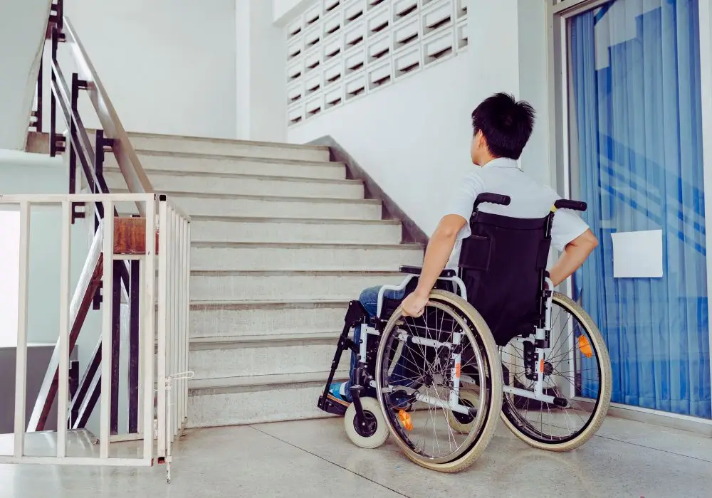 Jakie dofinansowanie do zamiany mieszkania dla niepełnosprawnych z PFRON można otrzymać? 1 Dostępne mieszkanie dla niepełnosprawnych – osoba poruszająca się na wózku inwalidzkim stoi przed schodami w budynku mieszkalnym, podkreślając bariery architektoniczne