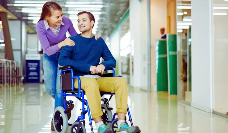 Asystent osoby niepełnosprawnej pomaga mężczyźnie poruszającemu się na wózku inwalidzkim w przestrzeni publicznej, okazując wsparcie i uważność.