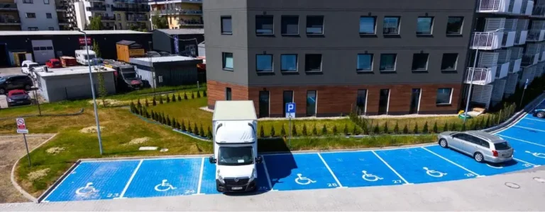 Zdjęcie do artykułu - miejsce parkingowe dla niepełnosprawnych. Obraz przedstawia parking z niebieskimi miejscami przeznaczonymi dla osób niepełnosprawnych. Widać kilka oznaczonych miejsc parkingowych z symbolami wózka inwalidzkiego. Na jednym z tych miejsc zaparkowany jest biały dostawczy samochód. W tle znajduje się ciemnoszary budynek mieszkalny z oknami oraz trawnikiem i żywopłotem. Na prawo od zaparkowanego samochodu widać zaparkowany szary sedan. W dalszym tle widoczne są inne budynki i samochody na drugim parkingu.