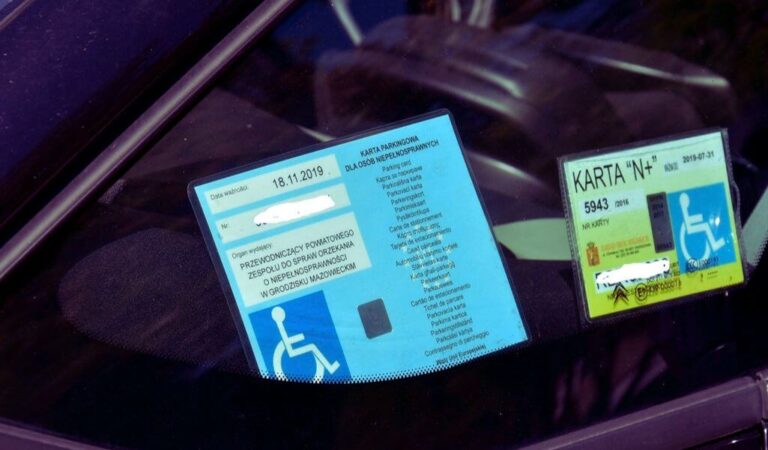 Zdjęcie przedstawia wnętrze samochodu, a dokładniej przednią szybę, na której znajdują się dwie karty parkingowe dla osób niepełnosprawnych. o Pierwsza karta (po lewej stronie): Jest koloru niebieskiego. Widnieje na niej data ważności. Pod datą jest miejsce na wpisanie numeru karty (wpisany numer jest zamazany). W dolnej części karty znajduje się symbol osoby na wózku inwalidzkim. Dodatkowo widoczny jest tekst w kilku językach, informujący o tym, że jest to karta parkingowa dla osób niepełnosprawnych. o Druga karta (po prawej stronie): Ma kolor jasnoniebieski i zielony. Na karcie widoczny jest numer oraz data ważności. Pod numerem znajduje się symbol osoby na wózku inwalidzkim. Obok symbolu widnieje tekst "Karta 'N+'". Na karcie są również wpisane dane, które są zamazane dla ochrony prywatności. Karty są umieszczone za przednią szybą samochodu, co wskazuje na to, że pojazd jest uprawniony do parkowania na miejscach dla osób niepełnosprawnych.