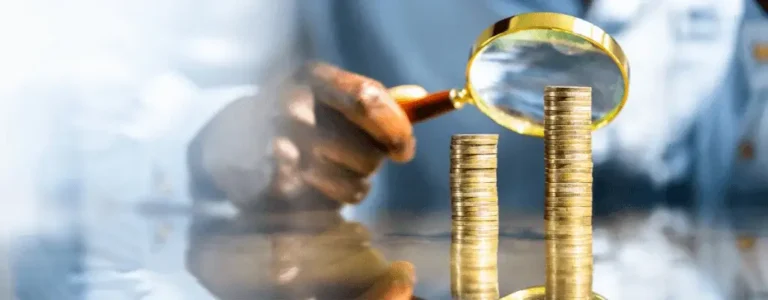 Stos monet oglądany przez lupę w ręku osoby, symbolizuje temat, jak wzrasta płaca minimalna.