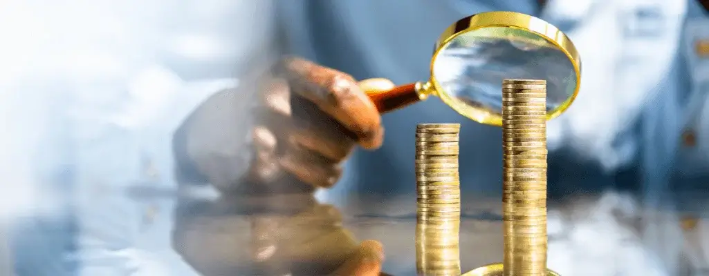 Stos monet oglądany przez lupę w ręku osoby, symbolizuje temat, jak wzrasta płaca minimalna.