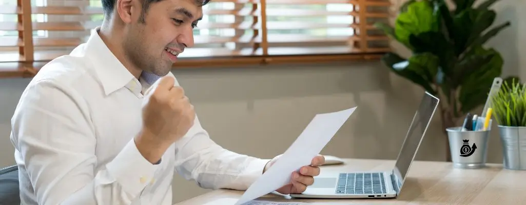 Obrazek wyróżniający do artykułu - podanie o podwyżkę. Na zdjęciu znajduje się mężczyzna siedzący przy biurku, ubrany w biały koszulę. Trzyma w ręku kartkę papieru i wyraża radość, co sugeruje jego podniesiona pięść. Przed nim na biurku znajduje się otwarty laptop. W tle widoczne są żaluzje na oknie oraz roślina doniczkowa, co wskazuje na to, że zdjęcie mogło zostać wykonane w biurze lub domowym gabinecie. Ogólne wrażenie zdjęcia jest pozytywne i profesjonalne.