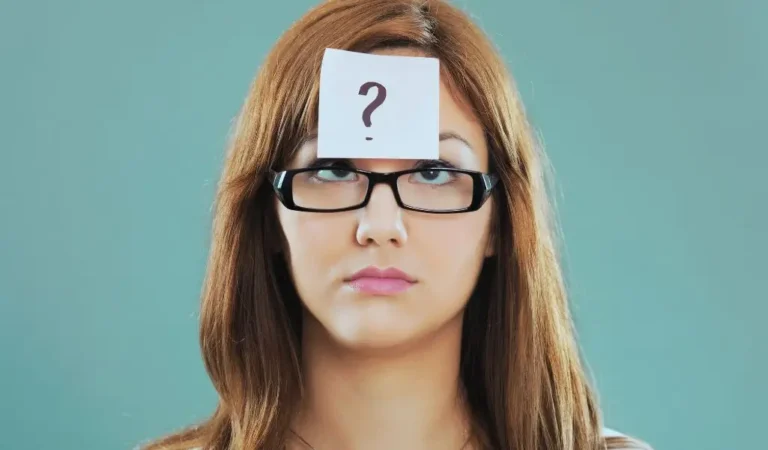 Zdjęcie do artykułu - słabe strony podczas rozmowy kwalifikacyjnej. Obraz przedstawia kobietę o jasnej karnacji i długich, prostych, brązowych włosach, która nosi okulary o prostokątnych, czarnych oprawkach. Kobieta patrzy w górę z neutralnym wyrazem twarzy. Na jej czole przyklejona jest kartka papieru z wydrukowanym dużym znakiem zapytania. Tło jest jednolite, w kolorze zielonkawo-niebieskim.