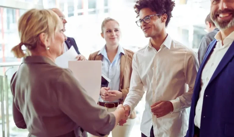 Obrazek wyróżniający do artykułu - Podziękowania za współpracę. Na zdjęciu widać grupę osób w formalnych strojach, które biorą udział w spotkaniu. Na pierwszym planie widoczna jest kobieta, która podaje rękę młodszemu mężczyźnie w białej koszuli. Oboje się uśmiechają, co sugeruje, że jest to przyjazne spotkanie. W tle znajdują się inne osoby, również ubrane formalnie. Jedna z kobiet trzyma dokumenty, a pozostali obserwują z uśmiechami na twarzach, co może świadczyć o oficjalnym zakończeniu spotkania lub wręczeniu podziękowań. Scena wydaje się być zorganizowana w profesjonalnym środowisku, prawdopodobnie w biurze lub sali konferencyjnej.