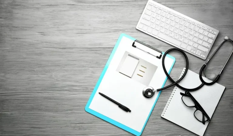 Obrazek wyróżniający do artykułu - lista chorób do orzeczenia o niepełnosprawności. Obraz przedstawia biurko z różnymi przedmiotami związanymi z pracą medyczną. Na szarym, drewnianym blacie znajduje się: Niebieska podkładka z klipsem, na której znajduje się notatnik i długopis. Białe, bezprzewodowe klawiatura komputerowa w górnej części obrazu. Stetoskop z czarnym przewodem i metalową głowicą, leżący obok notatnika. Czarna para okularów położona na notatniku w dolnej prawej części obrazu. Całość aranżacji sugeruje stanowisko pracy lekarza lub pracownika medycznego.