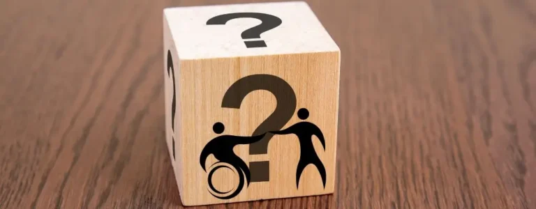 Zdjęcie w artykule - co daje orzeczenie o niepełnosprawności. Na obrazku widzimy drewniany sześcian ustawiony na powierzchni z drewna. Na jednym z boków sześcianu widnieje znak zapytania, a na innej stronie znajduje się grafika przedstawiająca osobę pomagającą innej osobie na wózku inwalidzkim. Ten obrazek symbolizuje pytania i wątpliwości związane z uzyskaniem orzeczenia o niepełnosprawności, sugerując, że może ono przynosić wsparcie i pomoc osobom niepełnosprawnym.