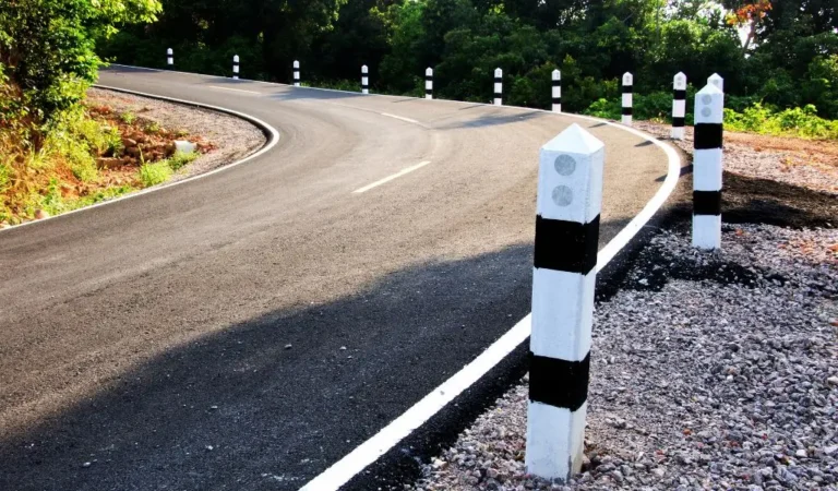 obrazek do artykułu - przelicznik mil na km. Obraz przedstawia asfaltową drogę, która łagodnie zakręca w lewo. Po obu stronach drogi znajdują się białe słupki z czarnymi paskami, które pomagają kierowcom ocenić odległość. W tle widoczne są drzewa i bujna roślinność, co sugeruje, że droga przebiega przez obszar wiejski lub leśny. Pobocze drogi jest pokryte żwirem.
