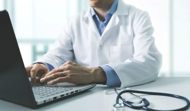 Obrazek wyróżniający do artykułu - zwolnienie lekarskie wstecz. Zdjęcie przedstawia lekarza siedzącego przy biurku, ubranego w biały fartuch medyczny. Jego ręce są na klawiaturze laptopa, na którym prawdopodobnie pracuje. Na biurku, obok laptopa, leży stetoskop. Tło jest rozmyte, z jasnym światłem wpadającym przez okna, co sugeruje, że scena ma miejsce w jasnym, profesjonalnym środowisku, prawdopodobnie w gabinecie lekarskim. Ogólny klimat zdjęcia to profesjonalizm i skupienie na pracy.
