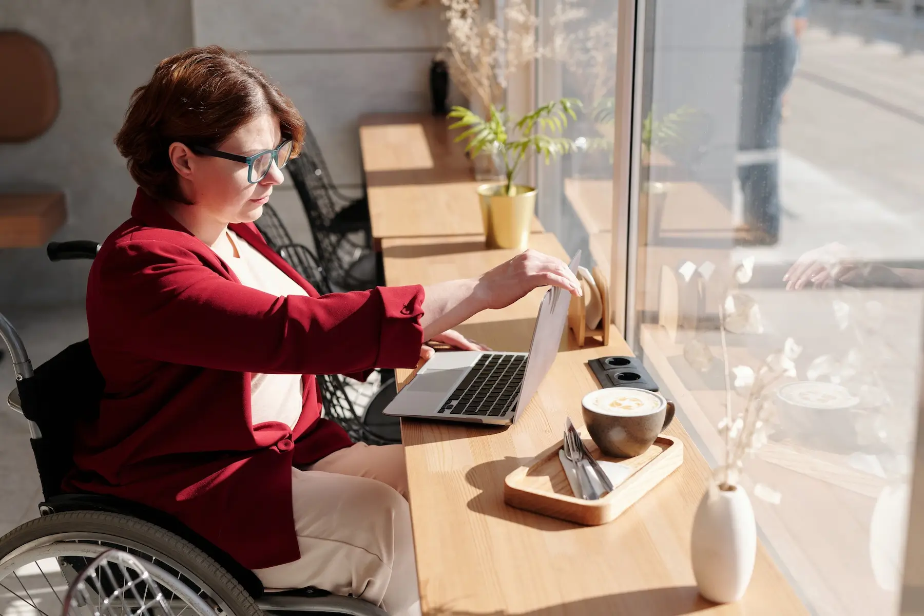 na zdjęciu kobieta z niepełnosprawnością na wózku. Przed komputerem, który stoi na długim biurku lub parapecie. Na drugim planie duże okno