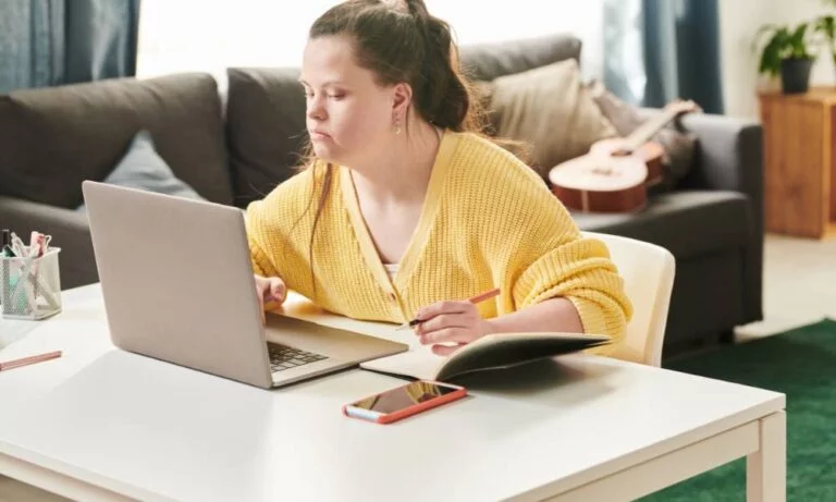 Jak otrzymać świadczenie wspierające w 2026? Wniosek WZON i ZUS, procedury krok po kroku 8 Zdjęcie główne artykułu o tytule „Jak otrzymać świadczenie wspierające? Poradnik krok po kroku” Na zdjęciu osoba z niepełnosprawnością. Siedzi ona przy stole, wyszukuje informacji w internecie na stojącym przed nią laptopie. Wykuje notatki, trzyma w dłoni ołówek.