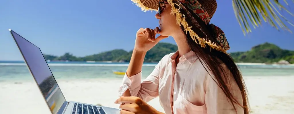 Zdjęcie w artykule - Freelancer kto to? Na zdjęciu zamyślona kobieta z laptopem na kolanach siedzi na plaży. Jej wzrok skierowany jest w dal. Zdjęcie sugeruje spokój i bezpieczeństwo.