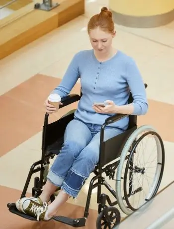 Zdjęcie przedstawia kobietę z niepełnosprawnością na wózku. W jednej dłoni trzyma papierowy kubek z kawą, w drugiej smartfon. Tło sugeruje, że znajduje się ona w galerii lub budynku użyteczności publicznej.