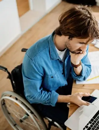 Zdjęcie przedstawiające mężczyznę, prawdopodobnie w pracy. Jest to osoba z niepełnosprawnością, siedząca na wózku przy biurku. Na biurku widoczny jest laptop, mężczyzna w dłoni trzyma smartfon. Mężczyzna wygląda na zastawiającego się nad czymś, brodę opiera na wewnętrznej części dłoni. 
