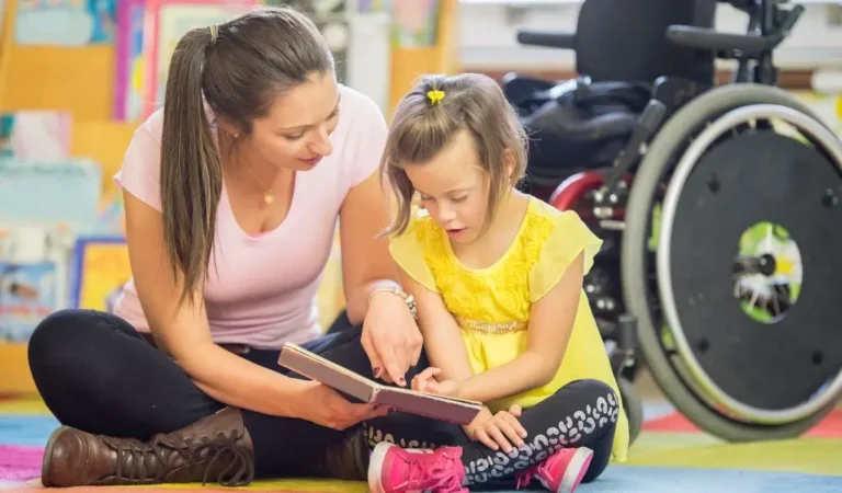 Obrazek wyróżniający w artykule - orzeczenie o potrzebie kształcenia specjalnego. Scenka w klasie. Na zdjęciu nauczycielka i uczennica siedzą na podłodze w klasie. Dziecko pokazuje coś w książce. W tle wózek inwalidzki. Widać przyjazną atmosferę i spokój oraz zainteresowanie ucznia.
