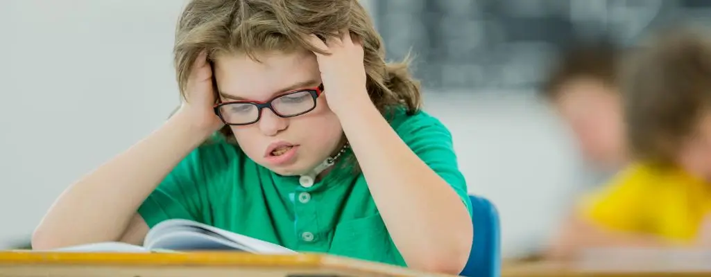 Zdjęcie w artykule - orzeczenie o potrzebie kształcenia specjalnego. Chłopiec z niepełnosprawnością w okularach podpiera bezradnie głowę i czyta coś w książce. W tle inne dzieci.