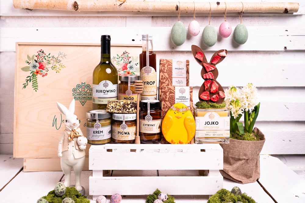 Zdjęcie przedstawiające wielkanocne prezenty dla pracowników. NA drewnianej skrzynce stoją różnorodne produkty, np. miody w butelkach i słoikach. Wielkanocny akcent to figurka zajączka.