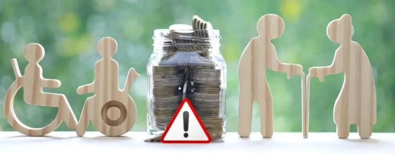 Drewniane figurki przedstawiające osoby z niepełnosprawnością i seniorów obok słoika z monetami; symbol ostrzeżenia wskazuje na problem finansowy — zabrano mi świadczenie pielęgnacyjne.
