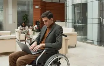 Obrazek osadzony w bloku o nazwie "znajdź pracownika". Przedstawia mężczyznę na wózku, który pisze na laptopie trzymanym na swoich kolanach. W tle obrazka panorama biurowa