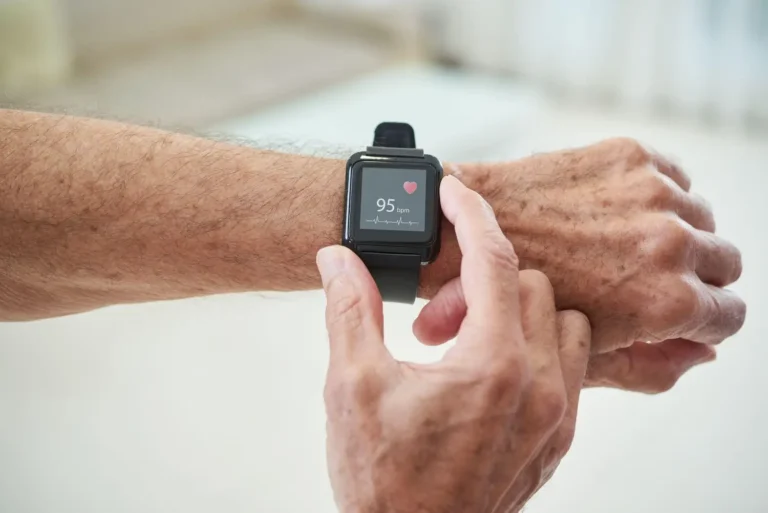 Na zdjęciu widzimy ręce starszej osoby z zegarkiem typu smartwatch na nadgarstku. Na ekranie zegarka wyświetlone są tętno o wartości 95 uderzeń na minutę i symbol serca. Tło jest jednolite i jasne.
