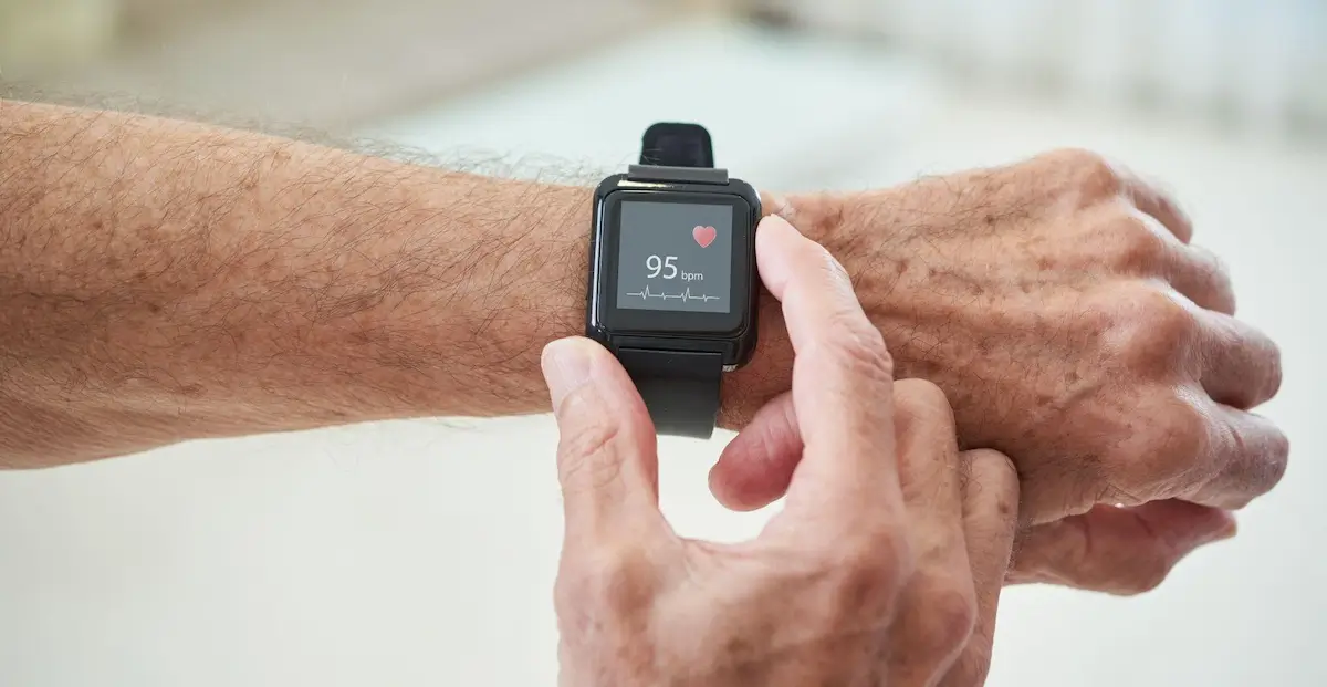  Na zdjęciu przedstawiona jest ręka starszej osoby noszącej czarny smartwatch, na którego ekranie wyświetlone są tętno o wartości 95 uderzeń na minutę i czerwone serduszko. Osoba dotyka ekranu smartwatcha drugą ręką. Tło jest neutralne, w jasnych odcieniach.