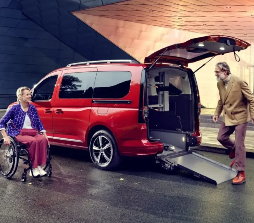 Obrazek wyróżniający do artykułu - dofinansowanie do samochodu dla osoby niepełnosprawnej. Na zdjęciu przedstawiono czerwony samochód kombi z otwartymi drzwiami bagażnika i zamontowaną na stałe platformą podnoszącą, która jest w trakcie opuszczania. Obok samochodu stoi kobieta na wózku inwalidzkim, uśmiechając się szeroko i patrząc w kierunku starszego mężczyzny, który obsługuje windę. Wyraz twarzy kobiety sugeruje zadowolenie i komfort, a cała scena wydaje się podkreślać niezależność i większą mobilność dzięki takiemu rozwiązaniu. Atmosfera jest pozytywna, a technologia wspomagająca dostępność pojazdu wydaje się ułatwiać codzienne życie.