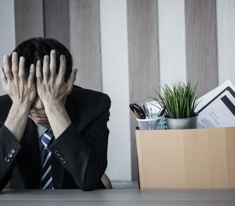 Obrazek wyróżniający w artykule - ile wynosi zasiłek dla bezrobotnych. Przedstawia mężczyznę w garniturze, który siedzi przy biurku z zakrytą twarzą rękoma. Wygląda na zrozpaczonego lub przytłoczonego. Na biurku przed nim znajduje się kartonowe pudełko, w którym leżą różne przedmioty biurowe takie jak długopisy i doniczka z zieloną rośliną, co może sugerować, że został zwolniony z pracy lub jest to scena po rozwiązaniu umowy o pracę.