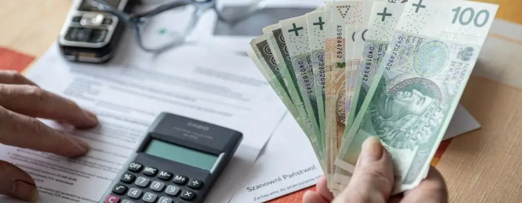 Zdjęcie w artykule - ile wynosi zasiłek dla bezrobotnych. Obrazek pokazuje ręce trzymające polskie banknoty nad biurkiem, na którym leży kalkulator i dokumenty. Wydaje się, że ktoś być może liczy oszczędności lub oblicza budżet domowy. Może to ilustrować moment otrzymania zasiłku dla bezrobotnych.