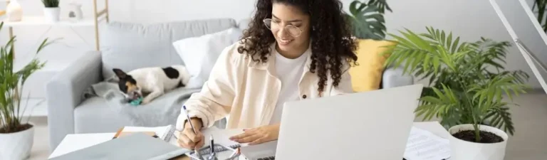 Obrazek wyróżniający do artykułu - Jakiej pracy nie znajdziesz na dzisiejszym rynku. Na obrazku widzimy młodą kobietę pracującą z domu. Siedzi przy biurku z laptopem, pisze na papierze i uśmiecha się lekko, sprawiając wrażenie zadowolonej. W tle widać jasne i przytulne wnętrze mieszkania oraz leżącego na kanapie psa, co może sugerować domową atmosferę i komfort pracy.