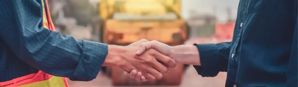 Obrazek wyróżniający do artykułu – kontrahent kto to. Zdjęcie pokazuje uścisk dłoni dwóch osób na tle rozmazanego miasteczka przemysłowego lub budowy. Jedna z postaci jest ubrana w niebieską kurtkę i odblaskowy kamizelkę, sugerując, że może być robotnikiem lub inżynierem. Druga osoba nosi ciemne ubranie, co może wskazywać na to, że jest wizytującym kontrahentem lub kierownikiem. Uścisk dłoni może symbolizować zakończenie negocjacji, zgodę lub rozpoczęcie współpracy.