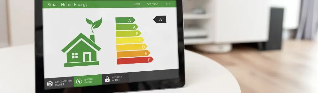 Obrazek wyróżniający do artykułu - oszczędzanie energii. Przedstawia tablet na białym stole, z ekranem wyświetlającym grafikę inteligentnego systemu zarządzania energią w domu. Ekran pokazuje ilustrację zielonego domu z liściem, obok której znajduje się pasek efektywności energetycznej z literami od A do F, gdzie A+ jest najwyżej i w kolorze zielonym, a F najniżej w kolorze czerwonym. Pod ilustracją domu znajdują się trzy ikony symbolizujące klimatyzację/ogrzewanie, oszczędność energii i alarm bezpieczeństwa. Obrazek wywołuje wrażenie nowoczesności i ekologicznego podejścia do zarządzania energią w domu.