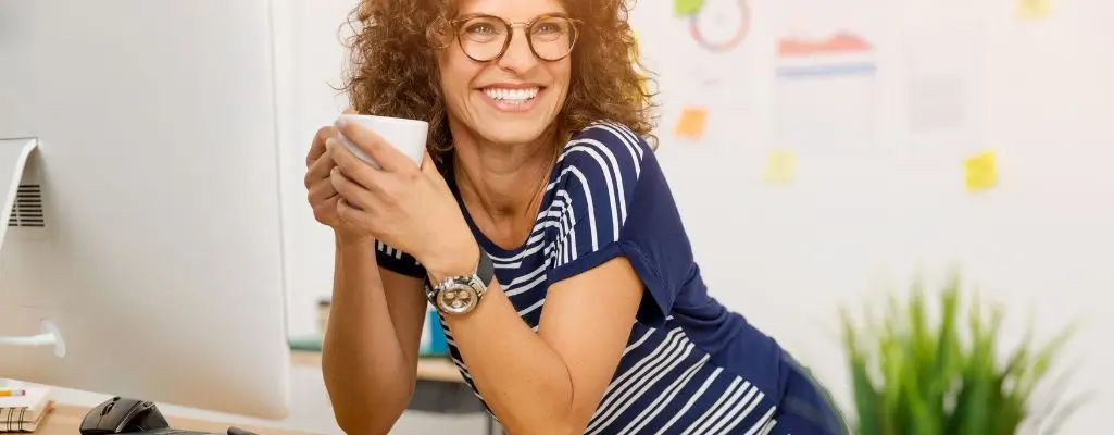 Obrazek wyróżniający do artykułu - przerwy w pracy. Przedstawia uśmiechniętą kobietę w okularach. Kobieta siedzi przy biurku, trzymając filiżankę, wydaje się zadowolona i zrelaksowana. Tło sugeruje biuro z różnymi notatkami w tle.
